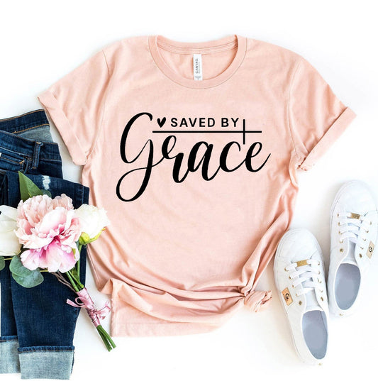 Pink t-shirt with 'Saved by Grace' text, jeans, sneakers, and flowers on a white background