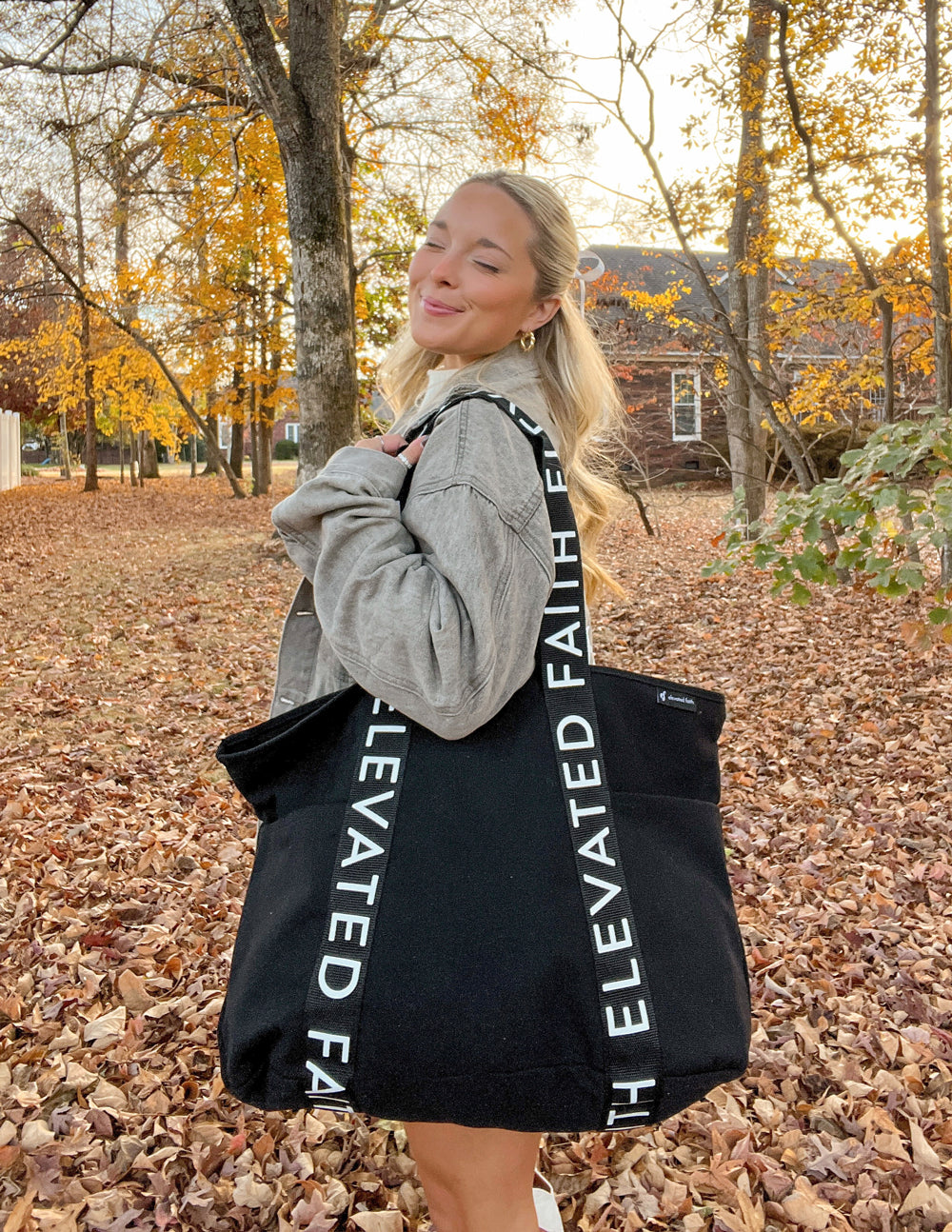 Woman holding a black tote bag with 'Elevated Faith' branding in an autumn setting.