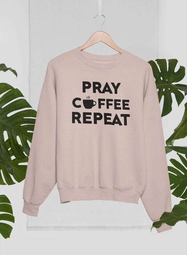 Beige sweatshirt with 'PRAY COFFEE REPEAT' text on a hanger with green leaves in the background