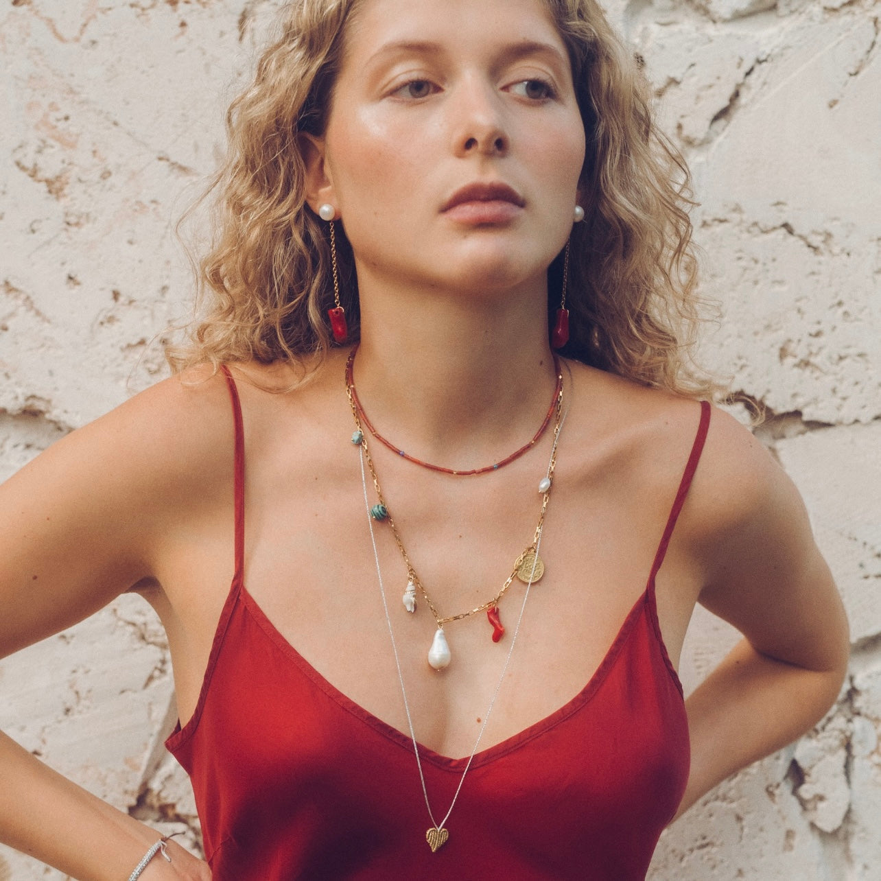 Woman wearing a red top and multiple necklaces with a stone wall background